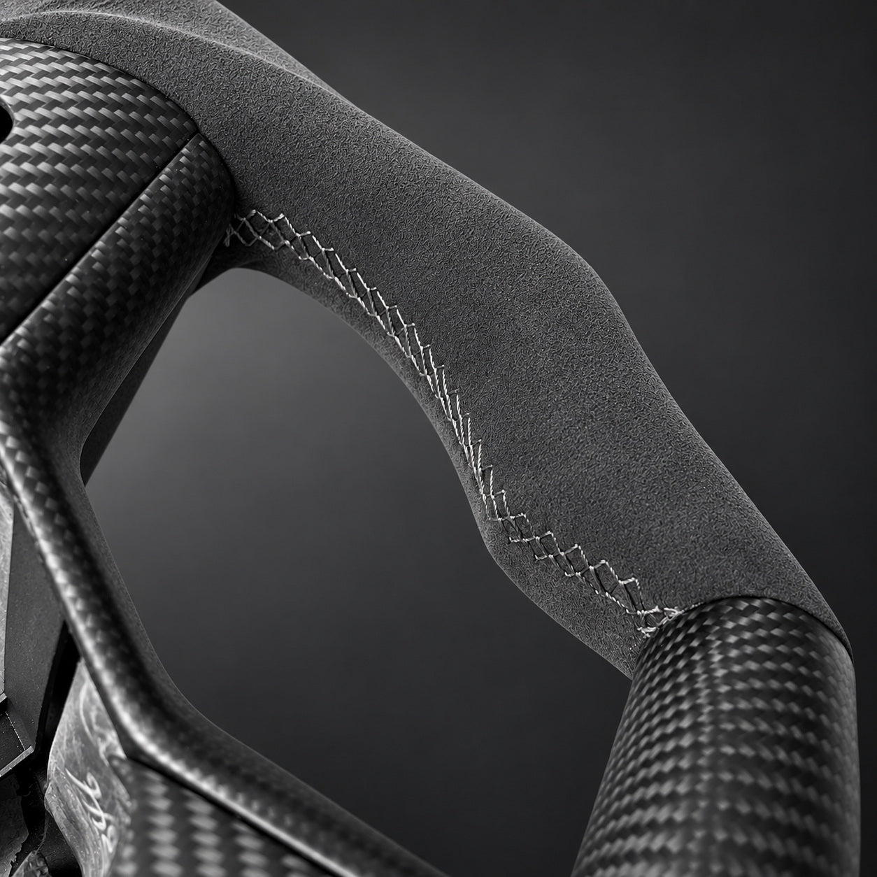 Grey Alcantara grip with grey cross stitching and adjacent woven carbon-fiber trim, shown in close detail.
